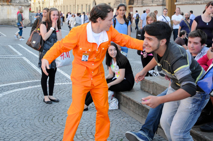 Die Fotografie zeigt eine künstlerische Intervention am Odeonsplatz. Ein Performer in orangenem Overall animiert eine Gruppe von Jugendlichen zur Teilnahme an der Aktion, die aber nicht im Bild zu sehen wird. Umstehend betrachten Passant*innen das Geschehen.