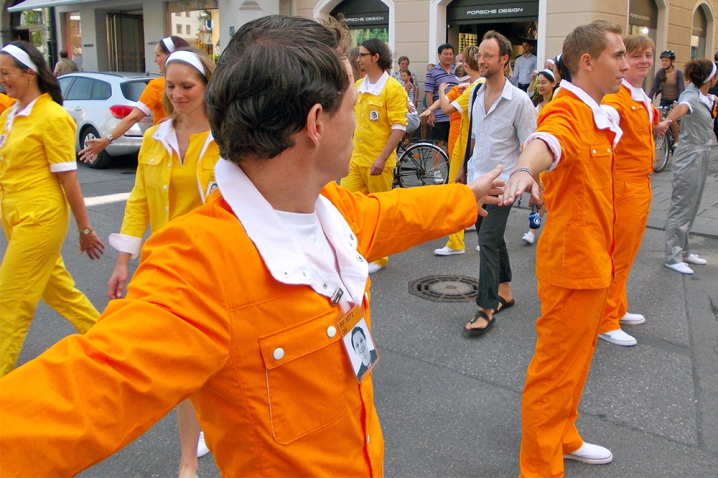 The image shows performers dressed in yellow, orange and silver overalls on Dienerstraße in the centre of Munich. Some performers hold hands and form two human chains with a corridor in the middle. Other performers and passers-by walk through this corridor.