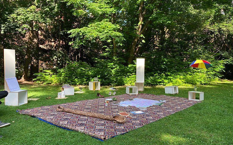 The photograph, taken in the Old Botanic Garden, shows a large carpet-like blanket lying on the grass. White wooden cubic seating objects are grouped around it. Various objects, such as maps and books, are spread out on the blanket. A colourful parasol can also be seen in the background.