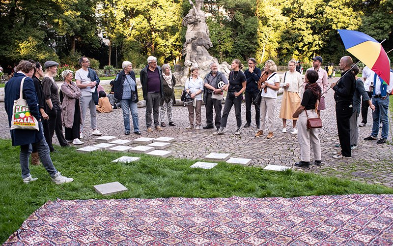 The photograph shows a group of people standing in front of the Neptune Fountain in the Old Botanical Garden. They appear to be taking part in a guided tour and the people are listening to a woman in the centre of the group. A man at the edge of the group is holding a colourful, open parasol over his shoulder.