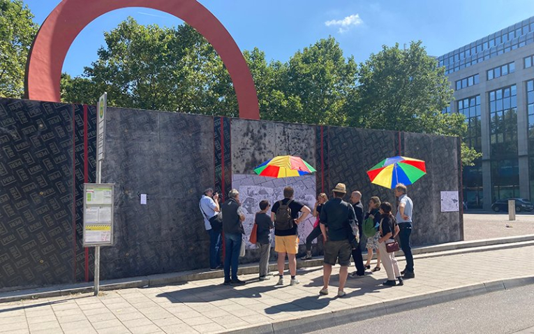 The photograph shows a group of people on the edge of the Old Botanical Gardens, apparently taking part in a guided tour. In the background is the sculpture ‘Der Ring “96” by Mauro Staccioli on the corner of Elisenstraße and Luisenstraße, which is surrounded by a dark temporary building site wall.