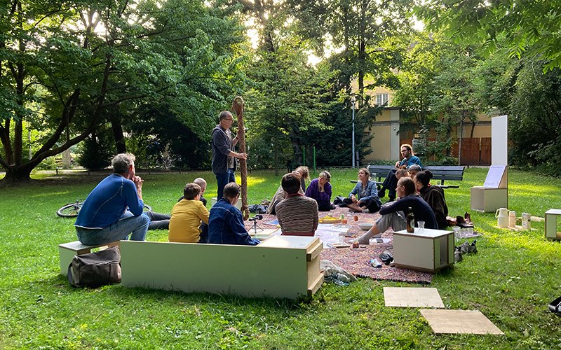 The photograph taken in the Old Botanical Garden shows a large carpet-like blanket lying on the grass. A group of people are sitting on the blanket and on white cubic seating objects grouped around it. They are listening to a man who is holding a large elongated object in his hands and explaining.