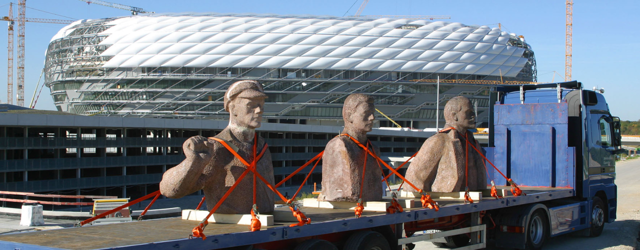 The photograph shows the travelling sculpture "Lenin on Tour" by Rudolf Herz in the foreground. Three granite busts of Lenin, taken from the Dresden Lenin Monument, are attached to a blue lorry with orange lashing straps. The unfinished Allianz Arena, which was under construction at the time and surrounded by cranes, can be seen in the background.