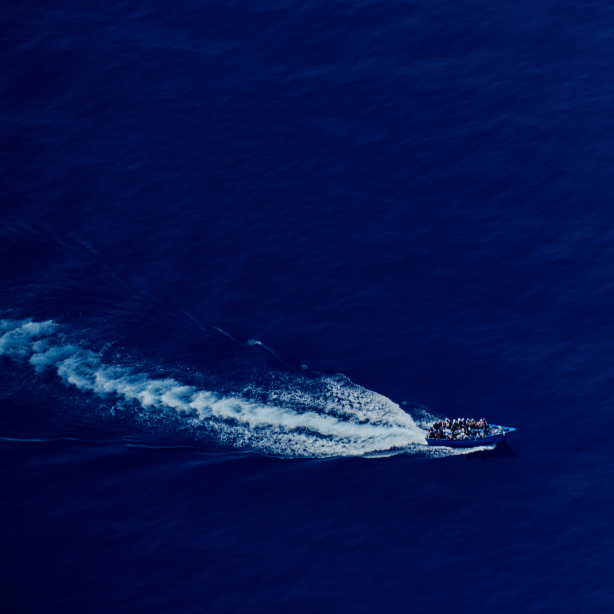 A bird's eye view shows a fully occupied boat with refugees in the open sea