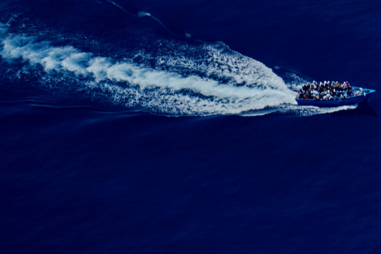 Zu sehen ist aus der Vogelperspektive ein voll besetztes Boot mit Flüchtlingen im offenen Meer