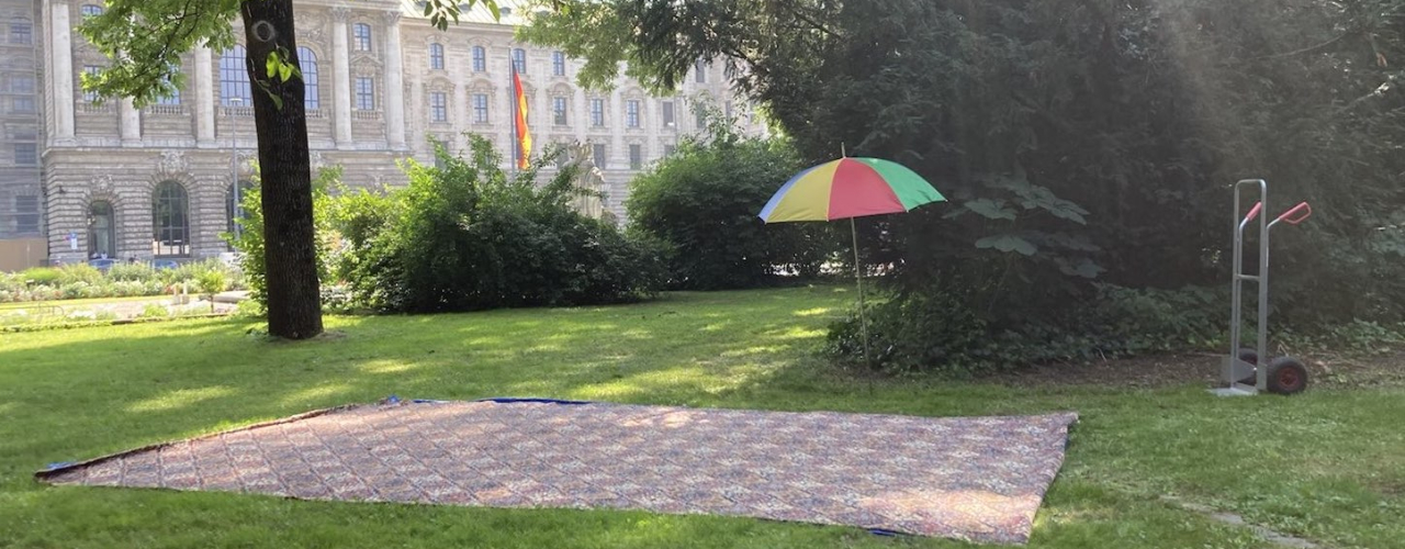 Photograph of a green space in the Old Botanical Garden. A large carpet-like rug has been laid out on the lawn, next to it is a sack barrow and a colourful parasol. The Palace of Justice can be seen in the background.