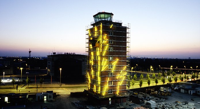 Fotografie des Kontrollturms des ehemaligen Flughafens München-Riem in der Abenddämmerung. Der eingezäunte Turm ist mit riesigen gelb leuchtenden Leuchtstäben übersäht, eine Lichtinstallation des Künstlers Chema Alvargonzalez.