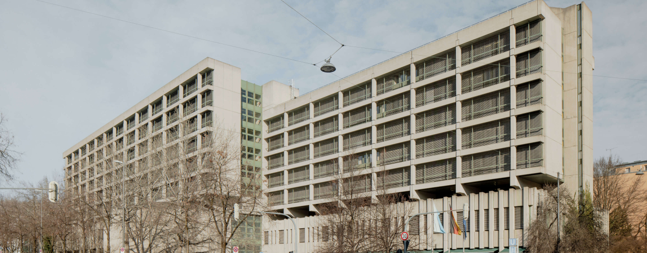 Photograph of the criminal justice centre on Nymphenburger Straße. You can see the grey exposed concrete building from the 1970s and the road junction in front of the building.