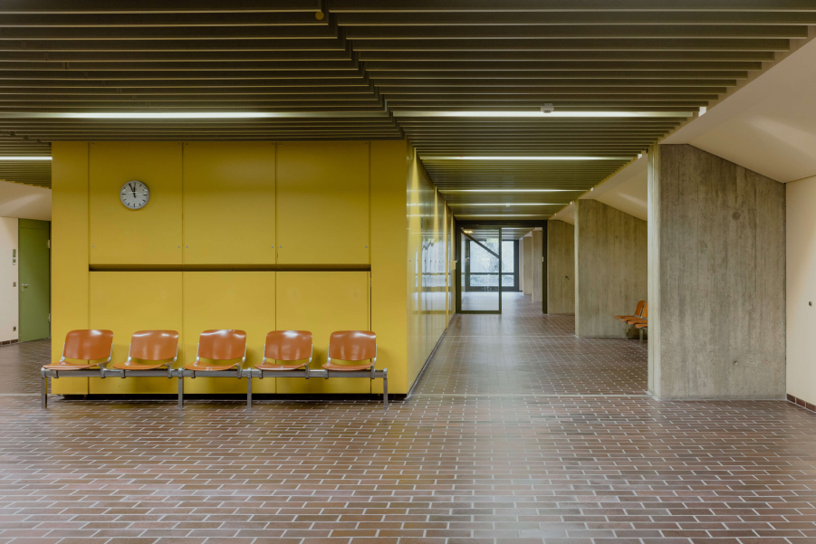 View of the interior of the Munich Criminal Justice Centre on Nymphenburger Strasse. The picture shows two illuminated corridors and a waiting area with a bench in front of a yellow panelled wall. The floor is covered with reddish-brown, brick-patterned tiles, while the walls feature a combination of grey exposed concrete and white plaster. The ceiling consists of horizontal louvres and linear strip lighting.