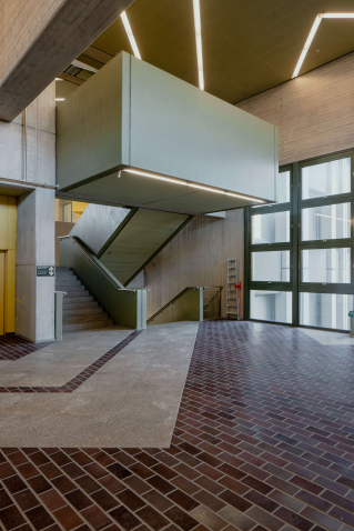 View of the interior of the criminal justice centre on Nymphenburger Straße. An open staircase can be seen, next to it a lift with a yellow door. The walls of the high interior rooms are made of grey exposed concrete, the floor is covered with rectangular tiles in various shades of brown.