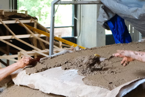 Herstellung des kompostierbaren SUVs, die lehmartige Masse wird auf ein mit Planen abgedecktes Holzgerüst aufgetragen.