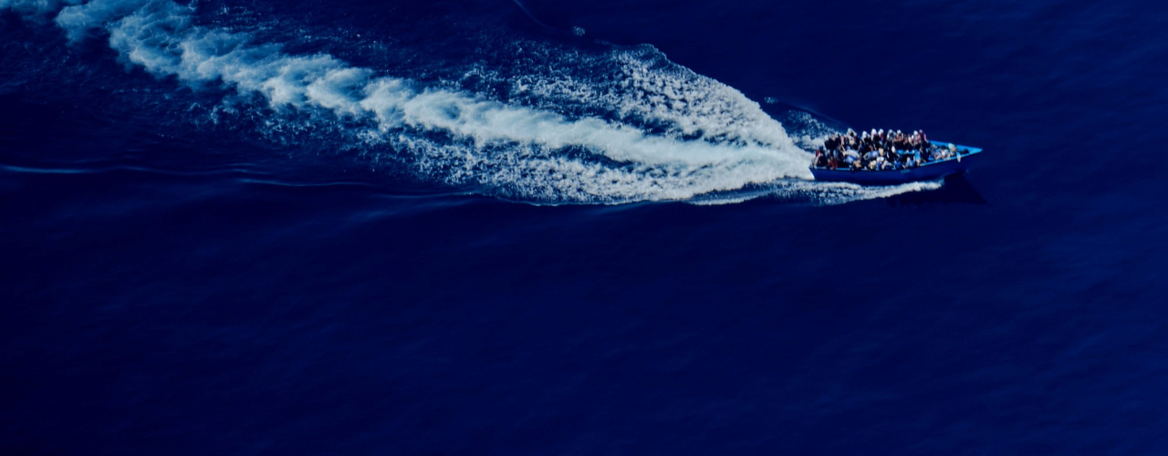 A bird's eye view shows a fully occupied boat with refugees in the open sea