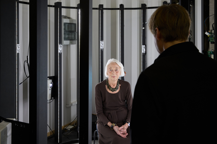 An elderly woman sits in a 3D scanner and looks into the camera. The artist Gregor Schneider can be seen from behind.