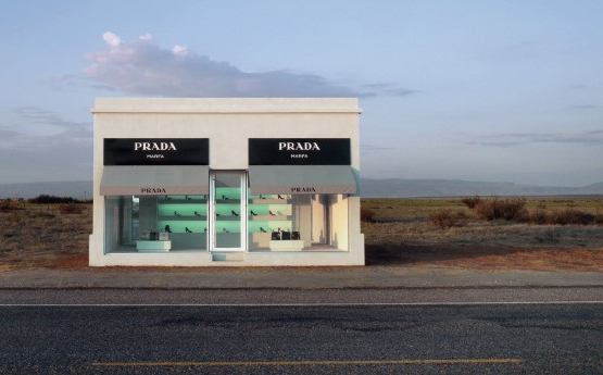 Das Bild zeigt eine einsame Prada-Boutique in einer Wüstenlandschaft, direkt an einer Straße, umgeben von leerer, trockener Natur.