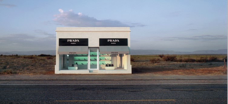 Das Bild zeigt eine einsame Prada-Boutique in einer Wüstenlandschaft, direkt an einer Straße, umgeben von leerer, trockener Natur.