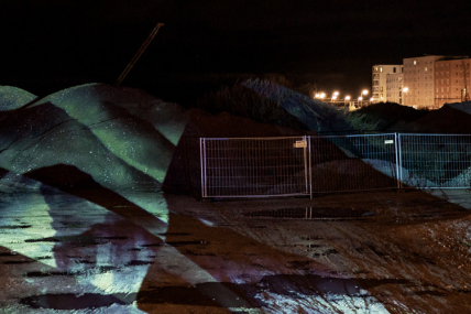 Das Bild zeigt eine Baustelle bei Nacht mit abgesperrten Erdhaufen, auf die eine abstrakte Lichtprojektion geworfen wird. Im Hintergrund sind beleuchtete Wohngebäude zu sehen.