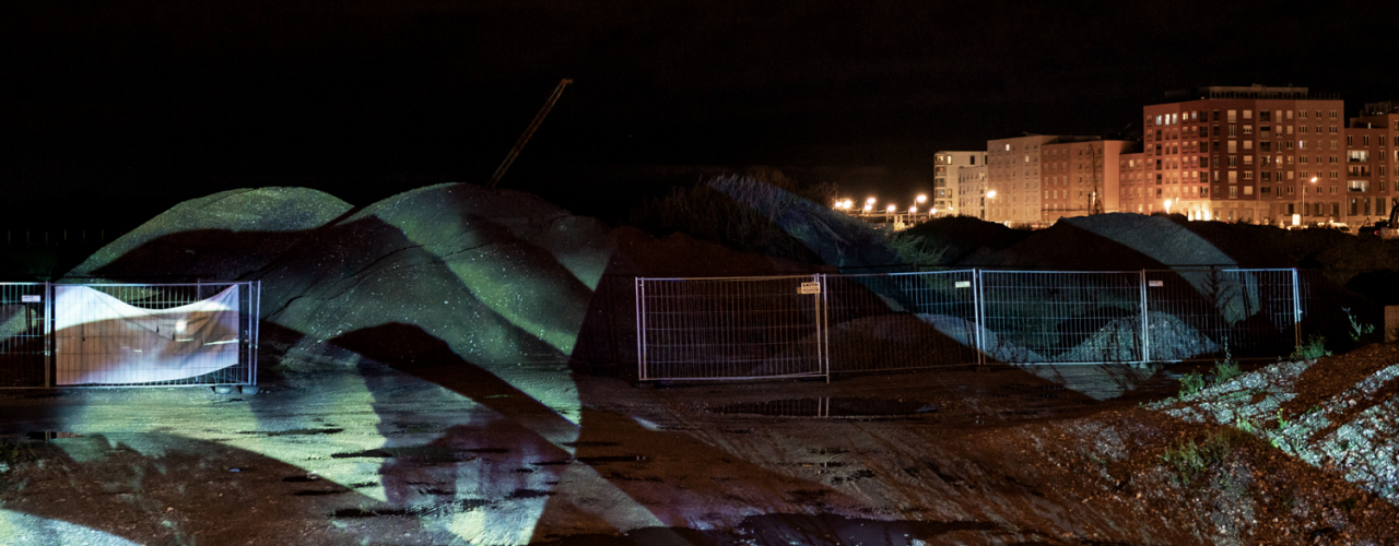 The image shows a construction site at night with fenced-off mounds of earth, illuminated by an abstract light projection. In the background, lit residential buildings are visible.