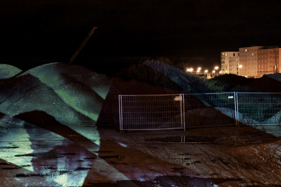 The image shows a construction site at night with fenced-off mounds of earth, illuminated by an abstract light projection. In the background, lit residential buildings are visible.