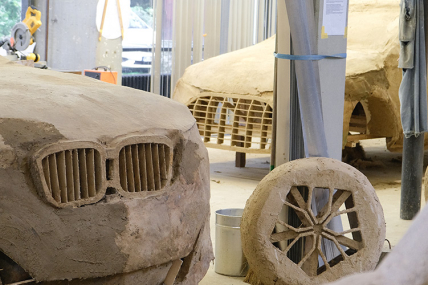 Blick in ein Arbeitsatelier. Darin sind drei Plastiken zu sehen, die SUVs in Originalgröße darstellen. Sie sind aus einem lehmartigen Stoff hergestellt.