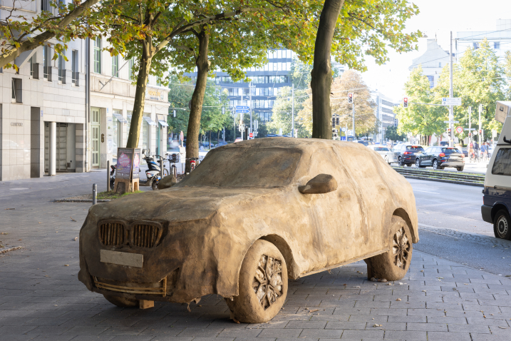 Ein aus Lehm geformter SUV steht an einer belebten Straßenecke vor einer geschäftigen Stadtlandschaft.