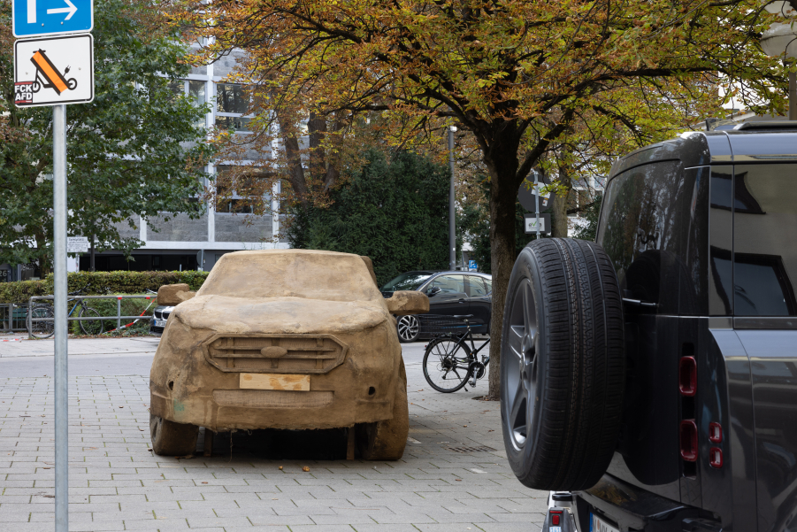 Das Bild zeigt einen aus Lehm geformten SUV, der in einer urbanen Umgebung auf einem Platz steht. Im Hintergrund sind Gebäude und Straßenlaternen sichtbar, im Vordergrund rechts ein echter schwarzer SUV.