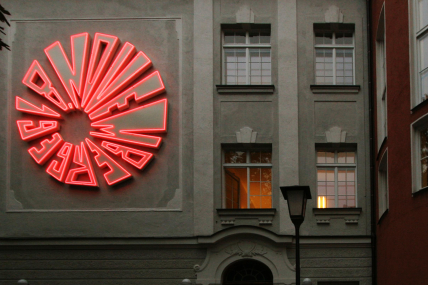 An einer Hauswandfassade ist in Höhe des 1. Stockwerks eine kreisrunde große Installation aus rot leuchtenden Neonröhren angebracht, die das Datum “8. November 1939“ ergibt.